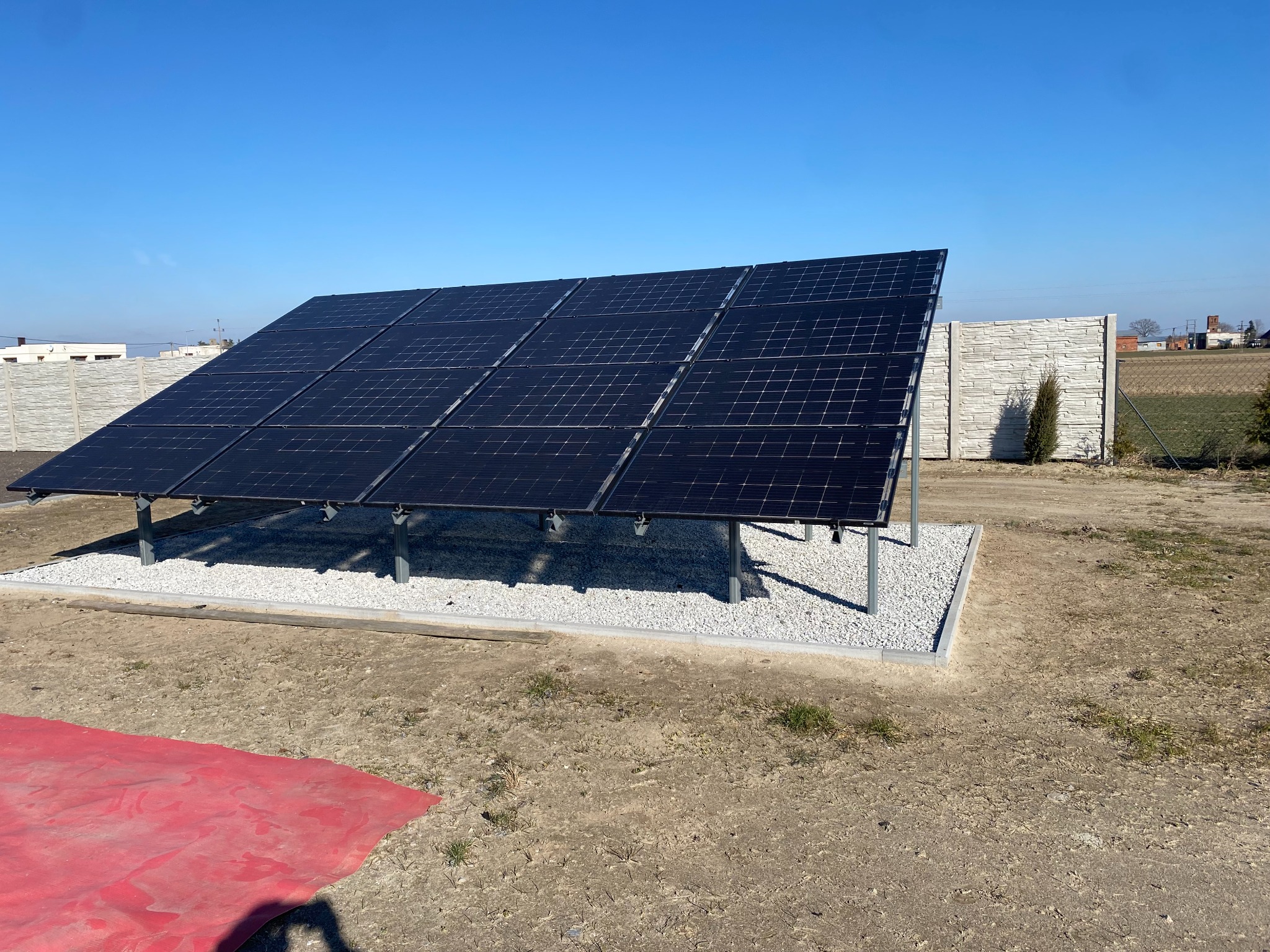 Panel słoneczny ustawiony na stelażu na gruncie z wysypanym białym żwirem, w tle ogrodzenie z betonowych elementów i zabudowania.