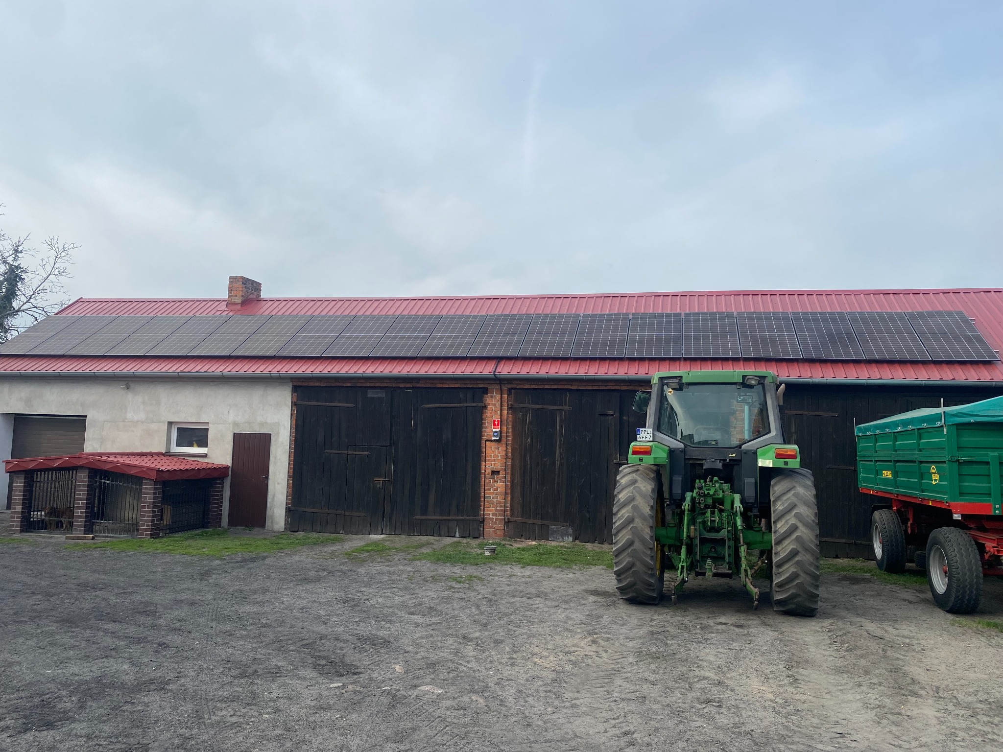 Instalacja fotowoltaiczna na dachu budynku gospodarczego z czerwonym dachem, na pierwszym planie traktor i przyczepa.