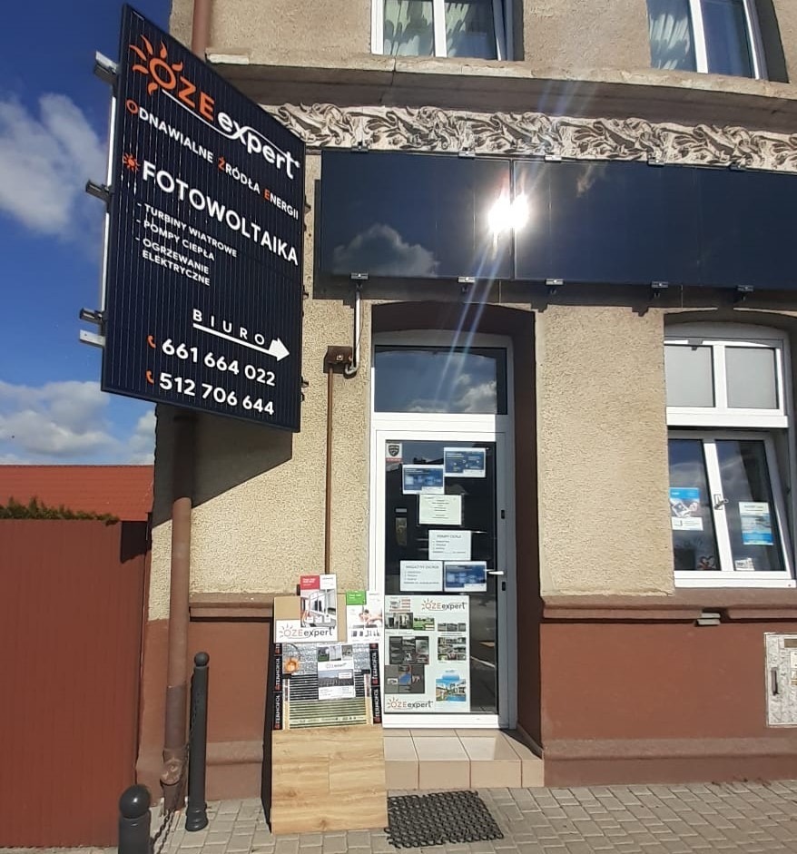 Fasada budynku z szyldem firmy OZE expert oferującej odnawialne źródła energii, w tym instalacje fotowoltaiczne, pompy ciepła i turbiny wiatrowe; nad wejściem zamontowane panele słoneczne...
