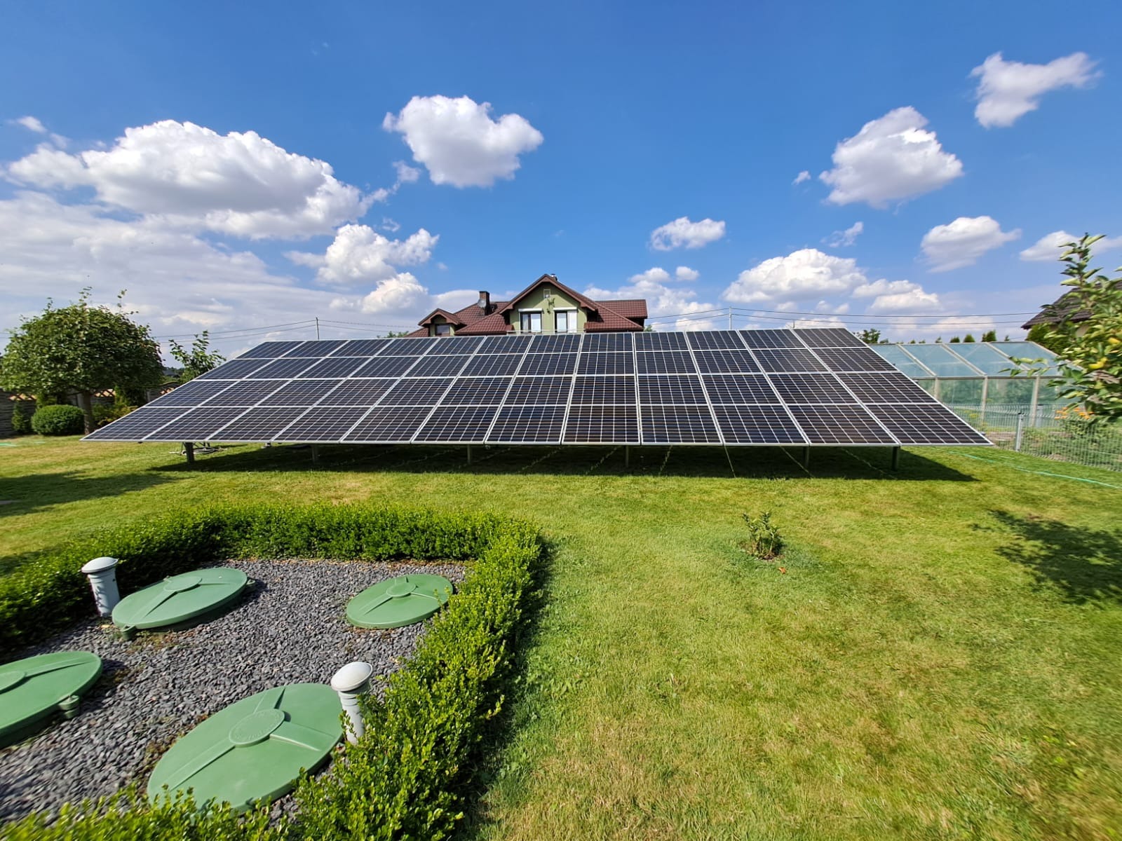 Instalacja paneli słonecznych na trawniku w Broniborze, moduły Risen RSM144-7-445 BMDG, falownik Sofar 11KTLX-G3, w tle dom jednorodzinny i szklarnia.