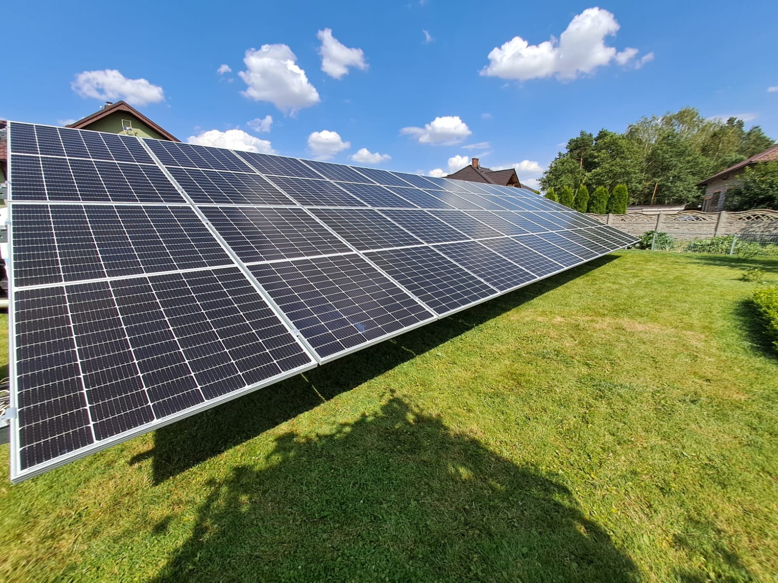 Instalacja paneli słonecznych na trawniku, widok z dołu, błękitne niebo z chmurami w tle.