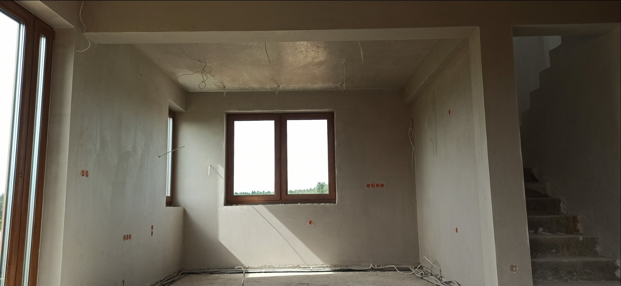 Wnętrze w surowym stanie po nałożeniu tynków, widoczne okna z drewnianą stolarką, wystające kable elektryczne i niedokończone schody.