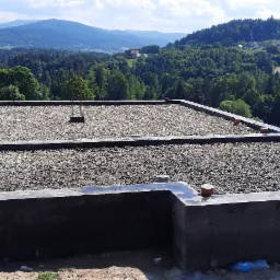 Wylany fundament z widoczną warstwą żwiru i wystającymi prętami zbrojeniowymi, na tle malowniczego krajobrazu g&oacute;rskiego.