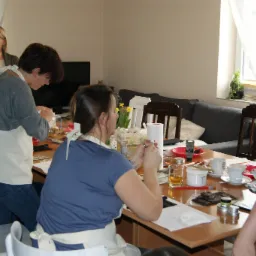 Uczestnicy warsztatów w fartuchach pracują przy stole z materiałami do rękodzieła, w tle okno z firanką i kanapa.