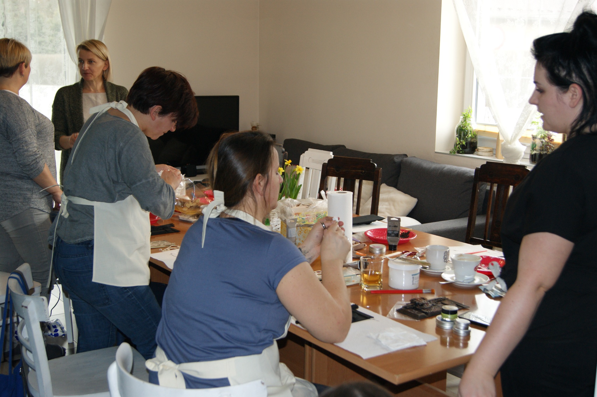 Uczestnicy warsztatów w fartuchach pracują przy stole z materiałami do rękodzieła, w tle okno z firanką i kanapa.