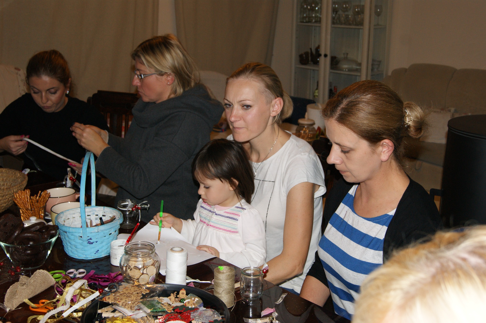 Grupa osób, w tym dziecko, uczestniczy w warsztatach rękodzielniczych przy stole zastawionym materiałami do tworzenia ozdób, z widocznym niebieskim koszykiem i słoikami z drewnianymi elementami.