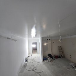 Pat-Bud - Białe, świeżo pomalowane ściany i sufit w remontowanym wnętrzu, z widocznymi narzędziami malarskimi i drabiną. Nowe, jednolite wykończenie.