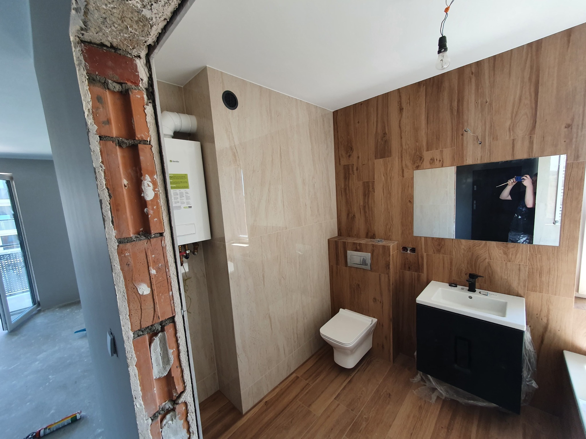 Wykończona łazienka z płytkami imitującymi drewno i beżowy marmur. Widoczna toaleta, umywalka z szafką i lustro. W tle kotłownia. W kadrze fragment ściany z cegły.