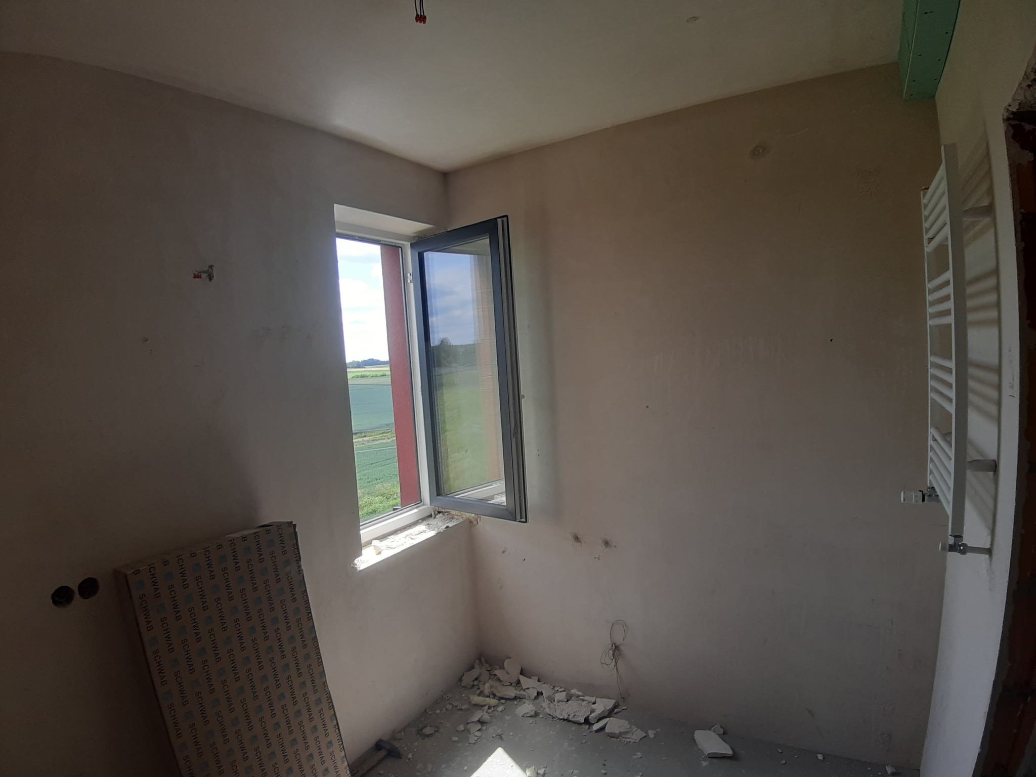 Pokój w trakcie remontu: ściany po szpachlowaniu, otwarte okno z widokiem na zieleń. Na podłodze resztki gruzu, przy ścianie płyty karton-gips. Grzejnik zamontowany.
