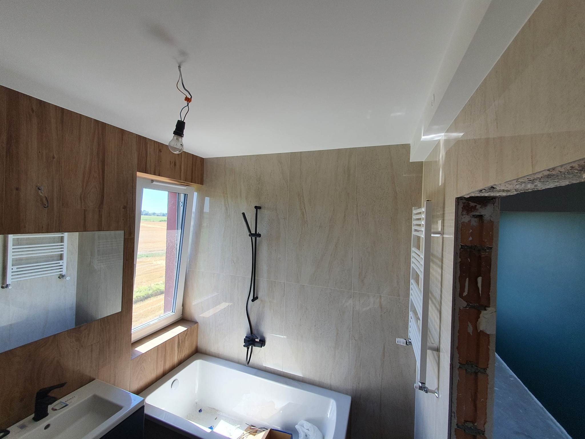 Jasna łazienka z beżowymi płytkami, białą wanną i umywalką. Widoczne okno z widokiem na pole, grzejnik drabinkowy i sufit z punktowym oświetleniem. Częściowo wykończone pomieszczenie.