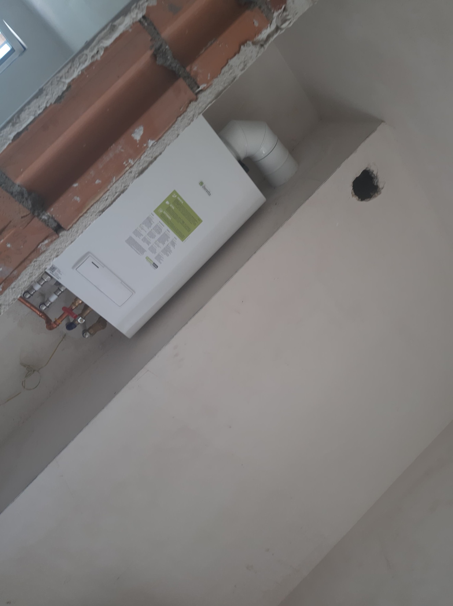 Zamontowany, biały piec gazowy marki Beretta, widoczny przyłączeniami rur miedzianych, na tle ściany z cegły i surowego tynku.