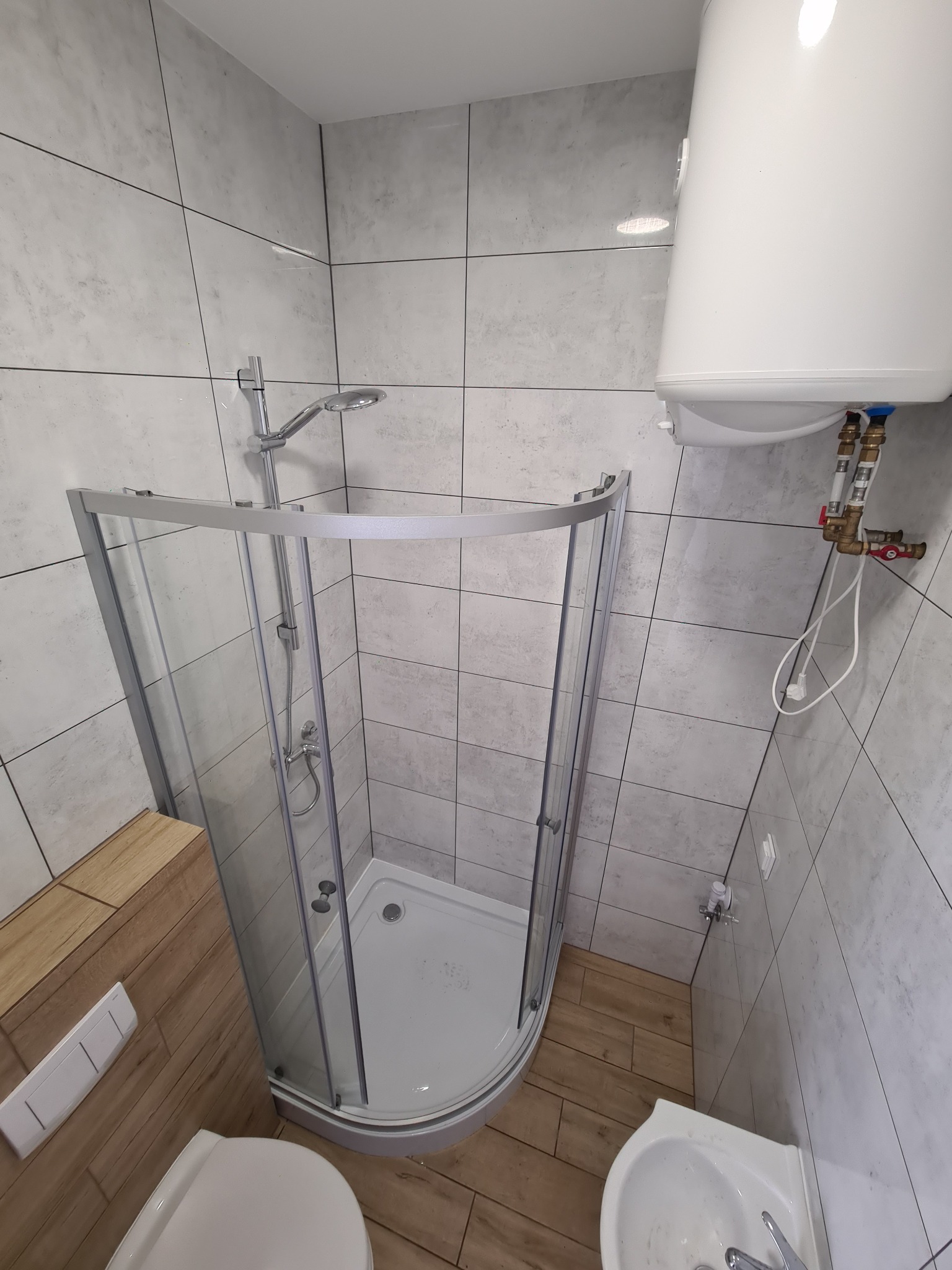 Nowoczesna kabina prysznicowa z brodzikiem, szara płytka imitująca beton, bojler elektryczny. Umywalka narożna i toaleta w jasnych barwach.