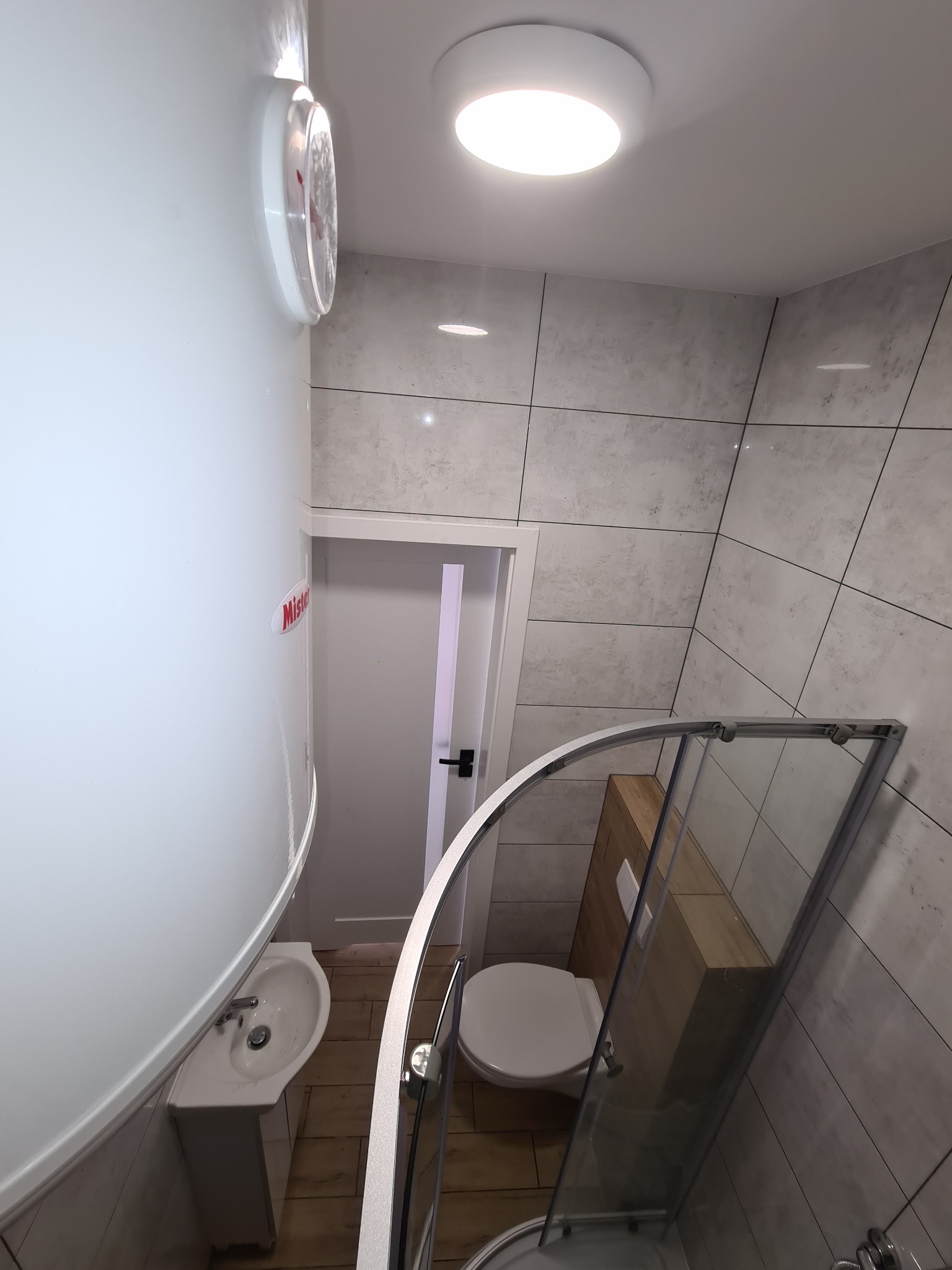 Wykończona łazienka w szaro-białej tonacji z kabiną prysznicową, umywalką i bojlerem. Widoczne płytki imitujące kamień i białe drzwi.