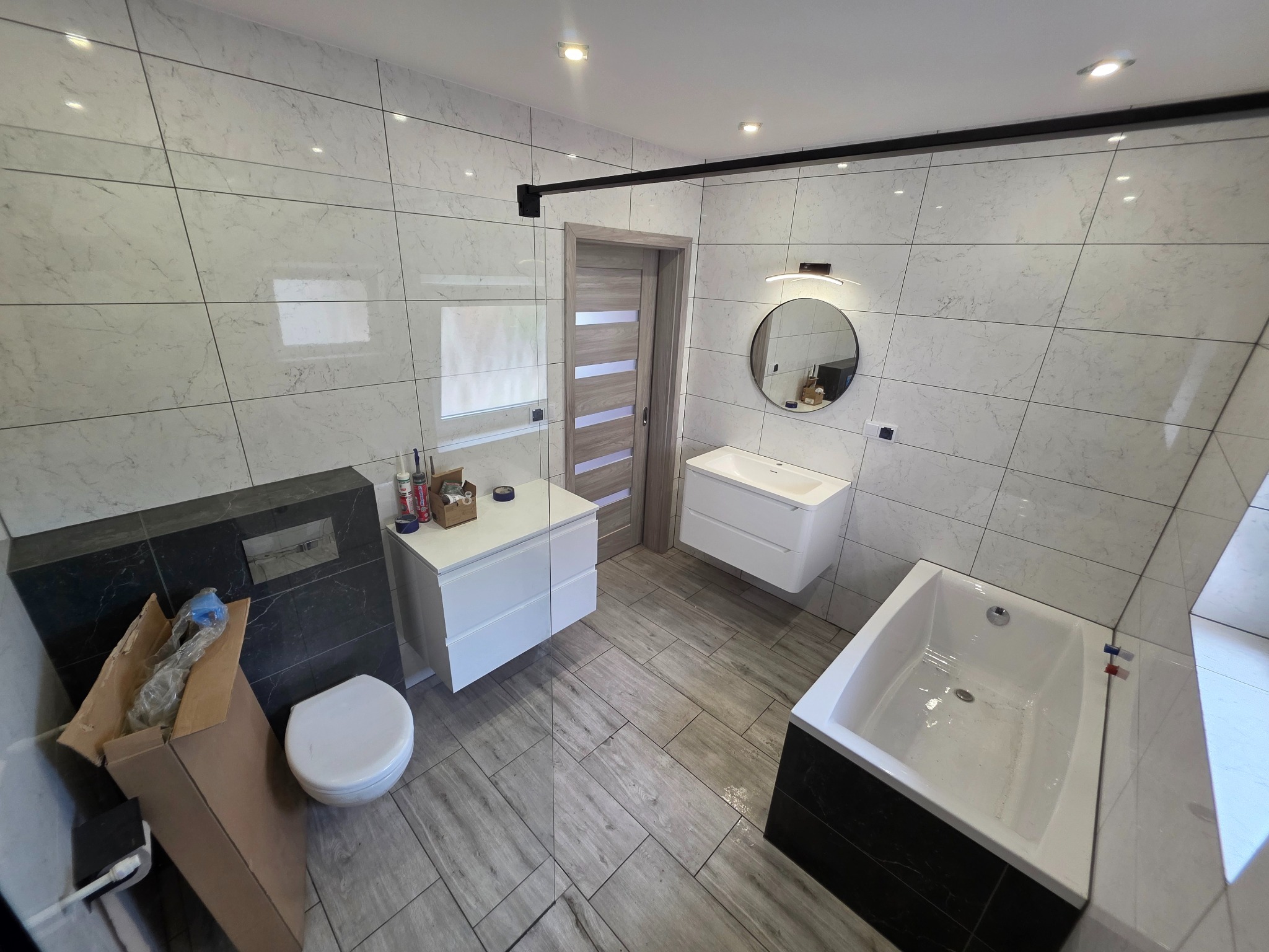 Nowoczesna łazienka z białymi płytkami imitującymi marmur, wiszącą armaturą, okrągłym lustrem i drewnopodobną podłogą. Widoczna kabina prysznicowa.