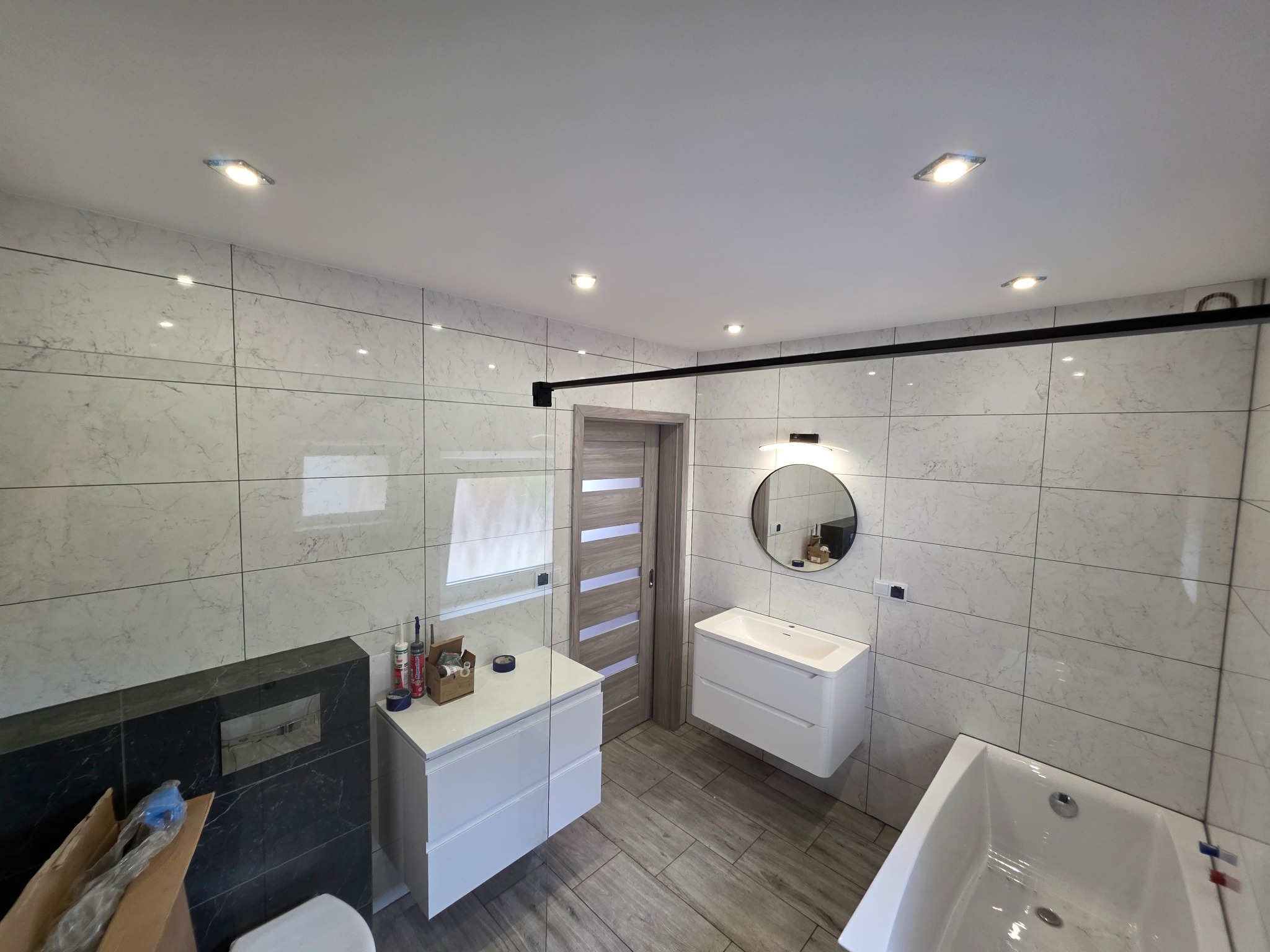 Nowoczesna łazienka z marmurowymi płytkami, białymi szafkami pod umywalki i czarną kabiną prysznicową. Widoczne oświetlenie punktowe i drewnopodobna podłoga.