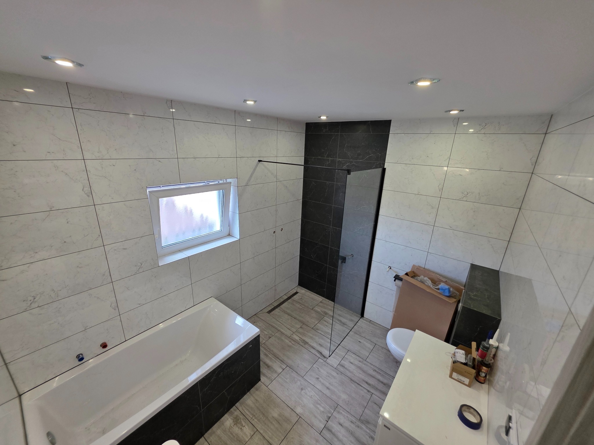 Wykończona łazienka z wanną, kabiną prysznicową i umywalką. Biało-czarne płytki na ścianach i podłodze. Widoczne oświetlenie sufitowe i okno.