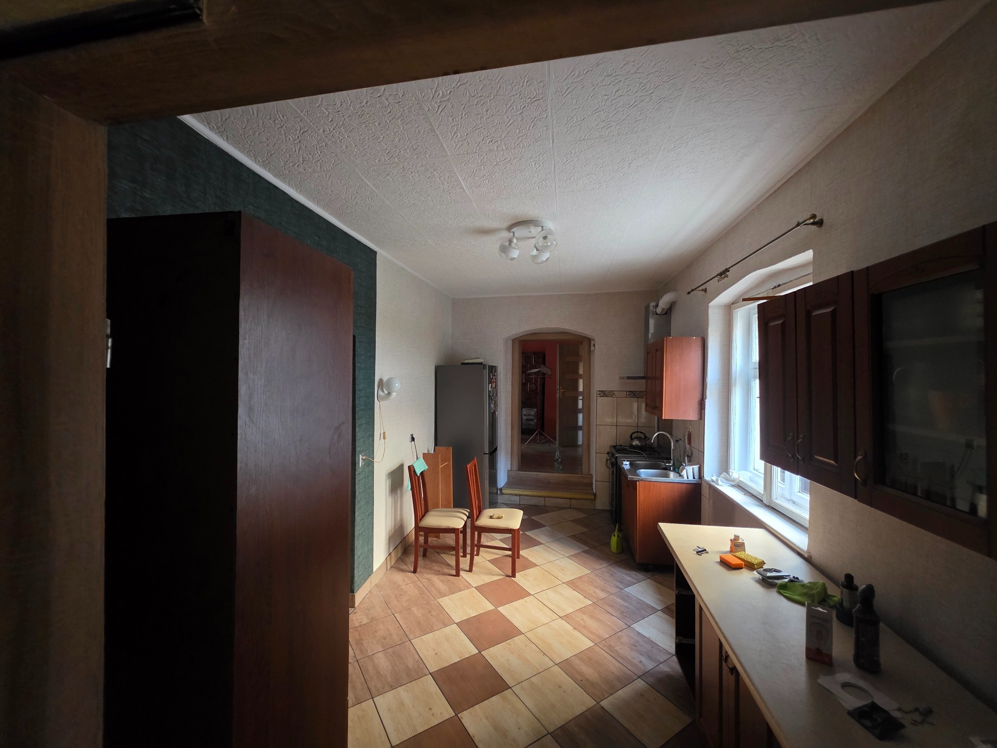 Wykończona kuchnia z brązowymi szafkami, wzorzystą podłogą i dekoracyjnym sufitem. Widoczne krzesła i sprzęt AGD. Całość w ciepłej tonacji, wnętrze po remoncie.