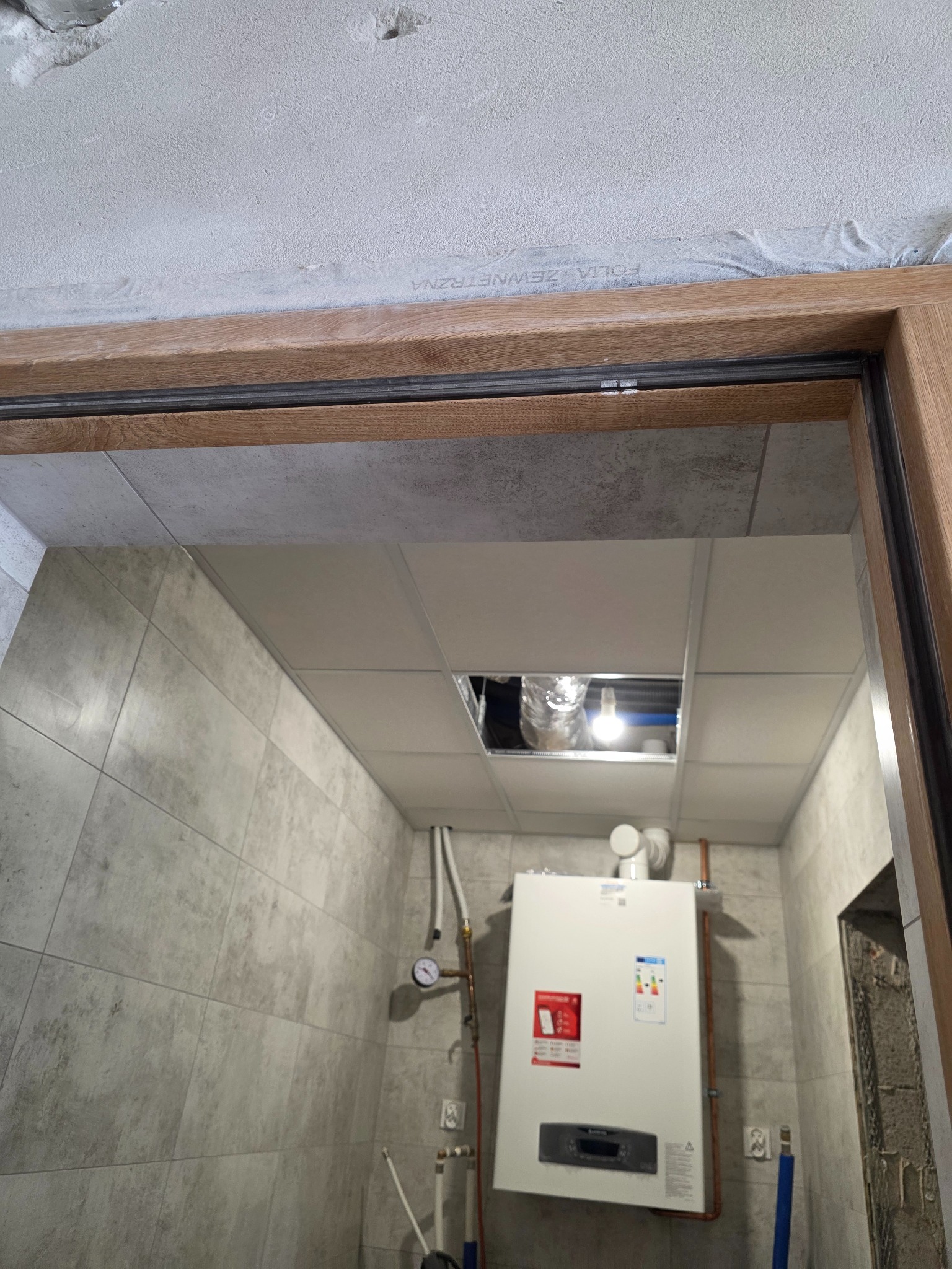 Wykończona łazienka z szarymi płytkami, widoczny bojler, sufit kasetonowy z otworem i drewniana ościeżnica drzwi. Ujęcie od dołu.