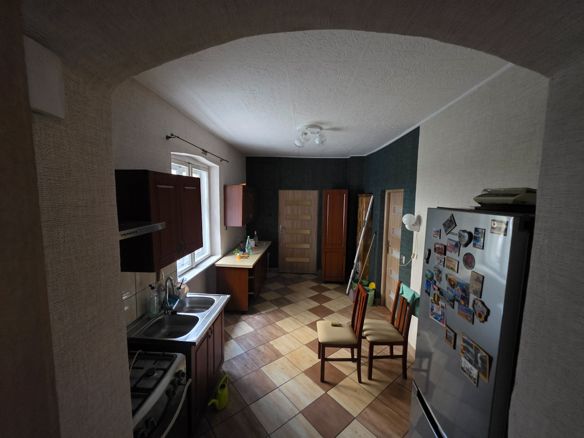 Widok na kuchnię z łukowym przejściem. Uwagę przyciągają brązowe płytki podłogowe ułożone w karo oraz liczne magnesy na lodówce po prawej stronie kadru.