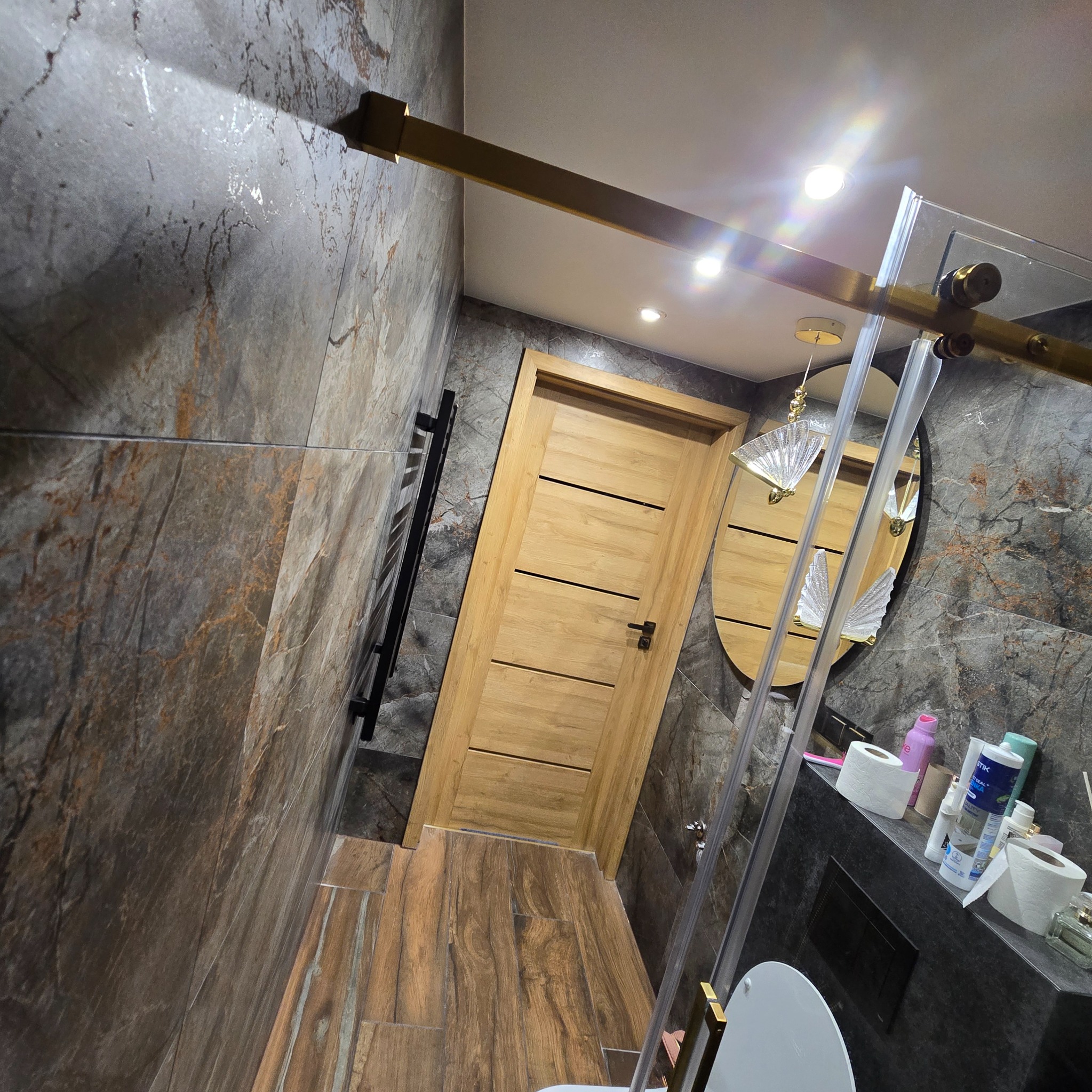 Stylowa łazienka z płytkami imitującymi marmur, drewnianą podłogą i drzwiami. Kabina prysznicowa ze złotymi akcentami i owalne lustro z dekoracyjnym oświetleniem.