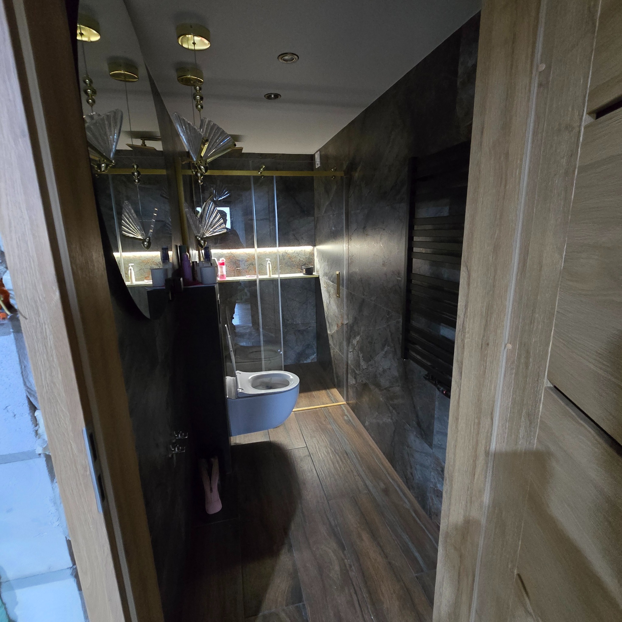 Elegancka łazienka z szarymi płytkami imitującymi marmur, drewnopodobną podłogą i złotymi akcentami oświetleniowymi. Widok przez drewniane drzwi.