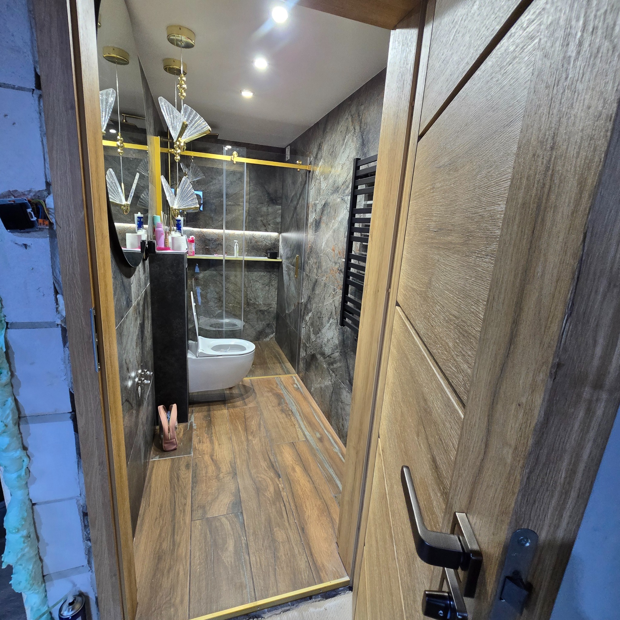 Nowoczesna łazienka z drewnopodobnymi płytkami, czarnymi akcentami i oświetleniem. Widok zza uchylonych drzwi, ukazujący kabinę prysznicową i toaletę.