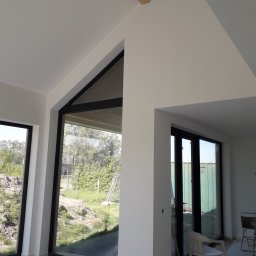 Firma Tynkarsko-Malarska DuDa - Wnętrze w trakcie prac wykończeniowych, świeżo pomalowane ściany na biało, duże okna z czarnymi ramami, widoczny fragment drewnianej belki stropowej i zabezpieczona podłoga.