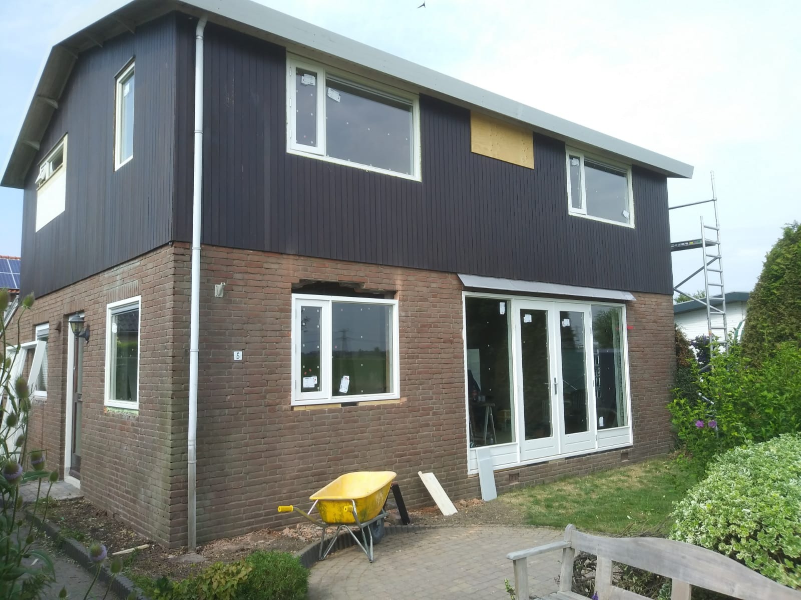 Dwupiętrowy dom z cegły i drewnianych paneli w trakcie renowacji, z żółtą taczką i rusztowaniem w tle.