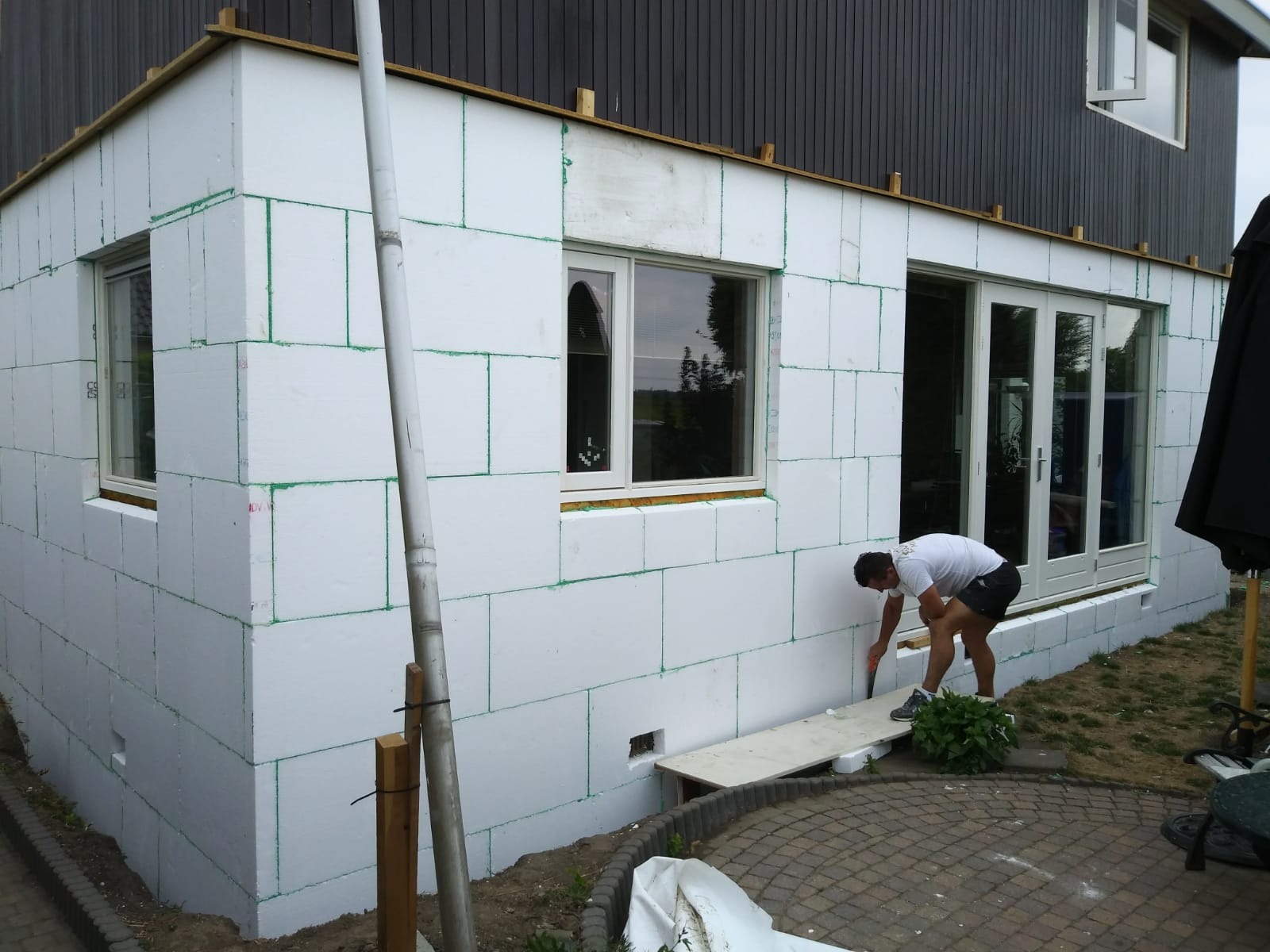 Prace związane z izolacją termiczną budynku: pracownik docina płytę styropianową na elewacji domu jednorodzinnego, widoczne okna i drzwi balkonowe.