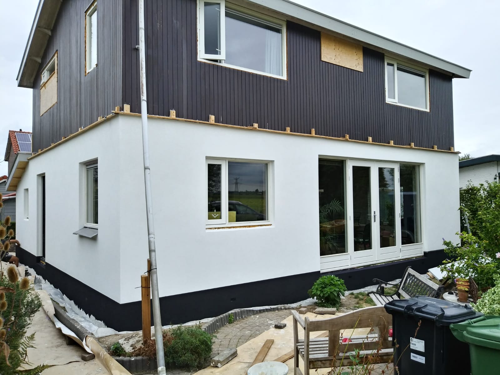 Dwukondygnacyjny dom z ocieploną, otynkowaną i pomalowaną na biało dolną częścią elewacji oraz ciemnobrązową, drewnianą górną częścią elewacji, z widocznymi drewnianymi elementami podtrzymującymi...