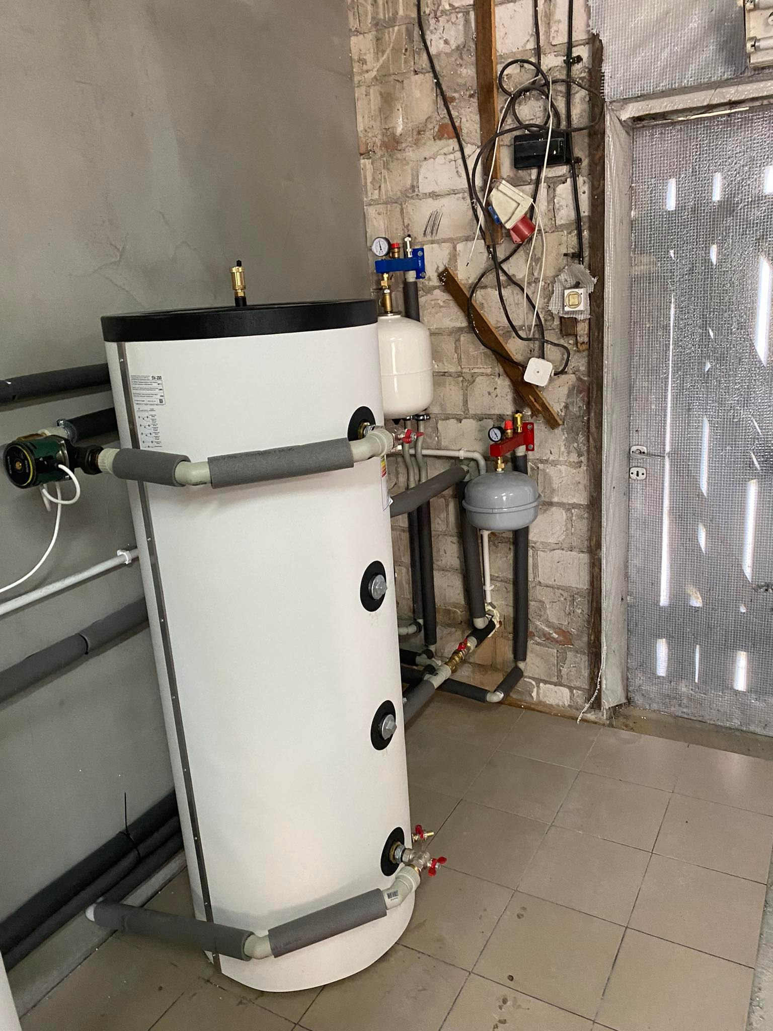 Bojler akumulacyjny z izolacją termiczną w kotłowni, widoczne rury i osprzęt, tło stanowią ściany z cegły i płytek.