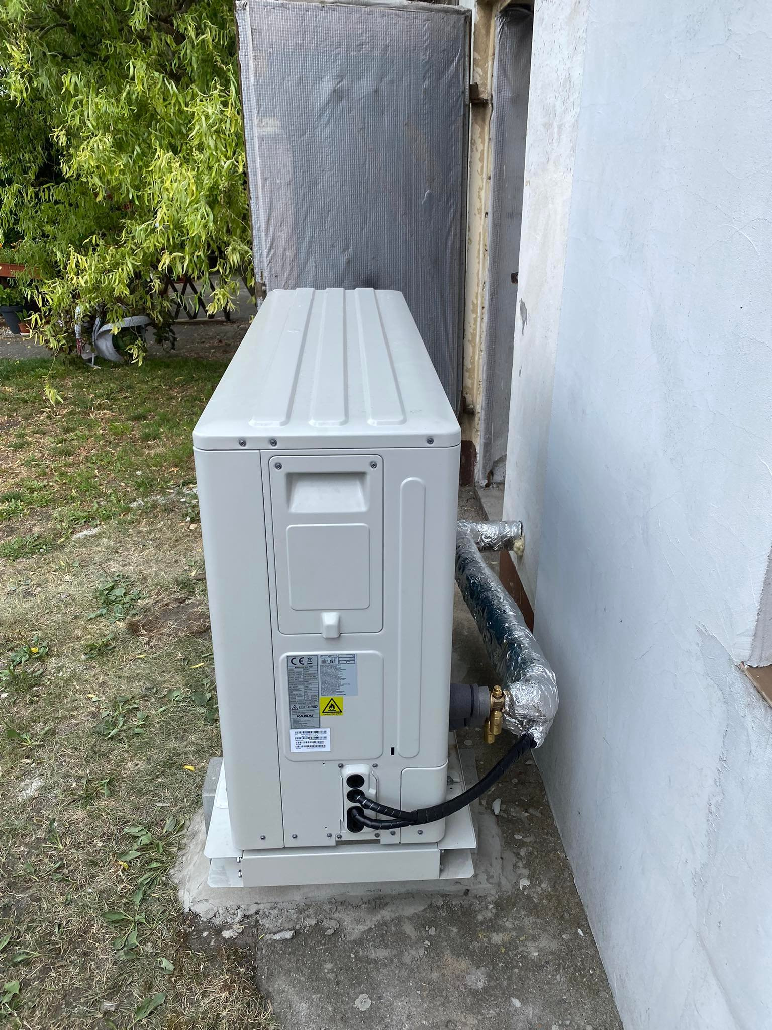 Zewnętrzna jednostka klimatyzacji typu split, marki KAISAI, zamontowana na betonowej podstawie przy białej ścianie budynku, z widocznymi rurami w izolacji i fragmentem zieleni w tle.