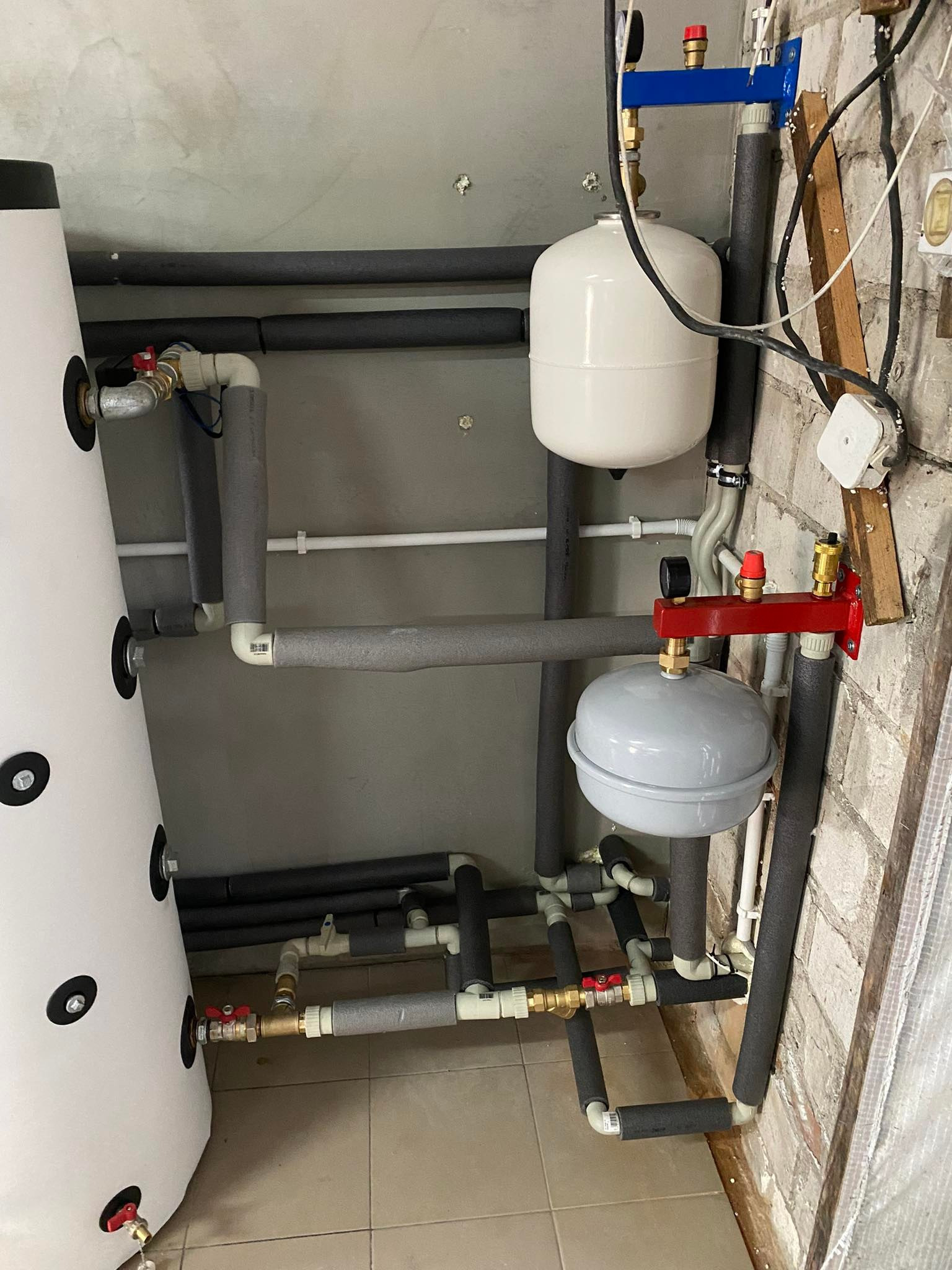 Instalacja z białym zbiornikiem buforowym, szarymi rurami z izolacją i czerwonym rozdzielaczem na tle ściany z cegły i szarej ściany.