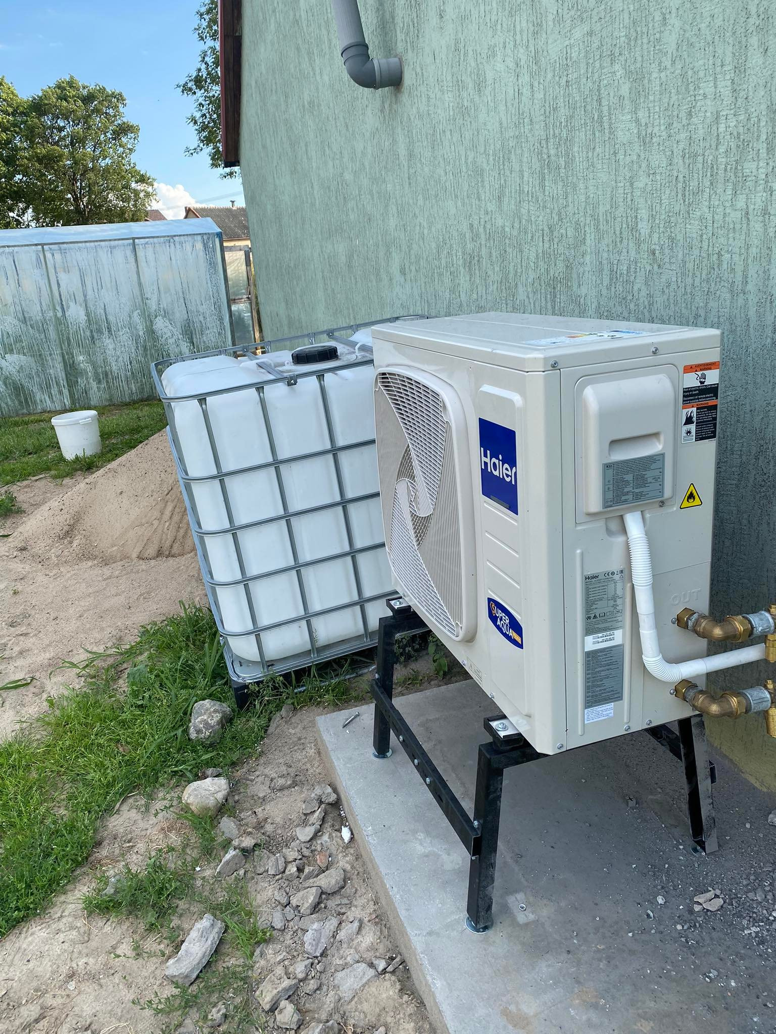 Zewnętrzna jednostka pompy ciepła Haier, zamontowana na czarnej metalowej konstrukcji, obok zbiornika na wodę, na tle ściany budynku o zielonkawej barwie.