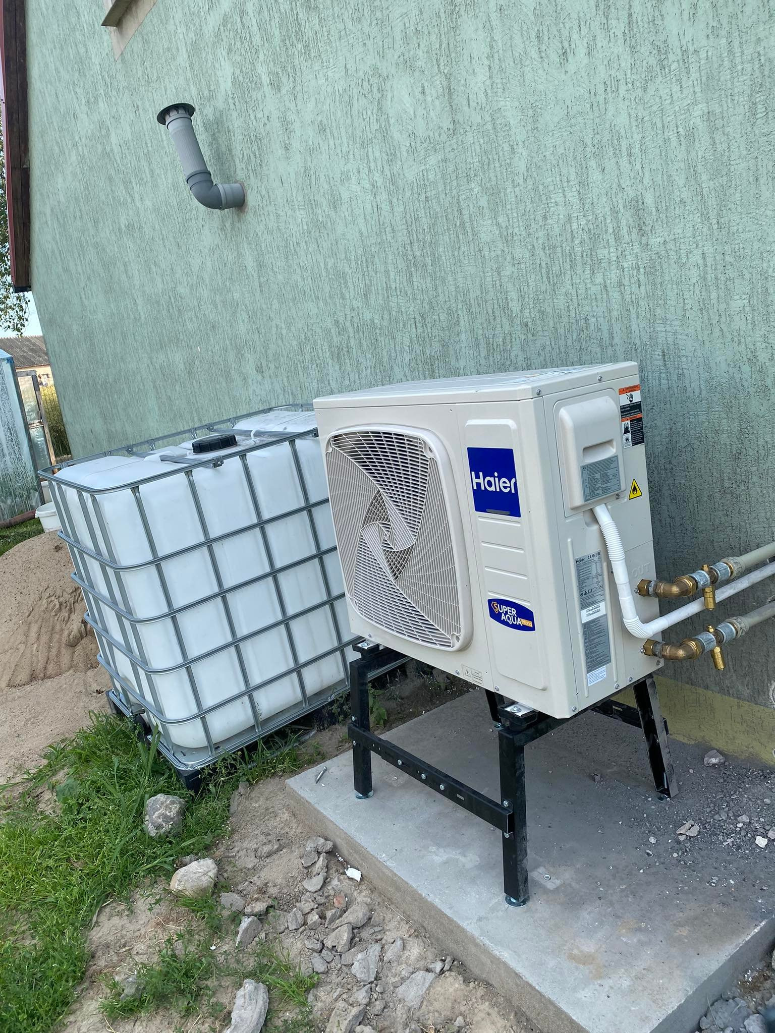 Biała, zewnętrzna jednostka klimatyzacyjna Haier zamontowana na czarnej, metalowej podstawie, z podłączonymi miedzianymi rurami, stoi obok białego zbiornika IBC na tle jasnej, teksturowanej ściany...