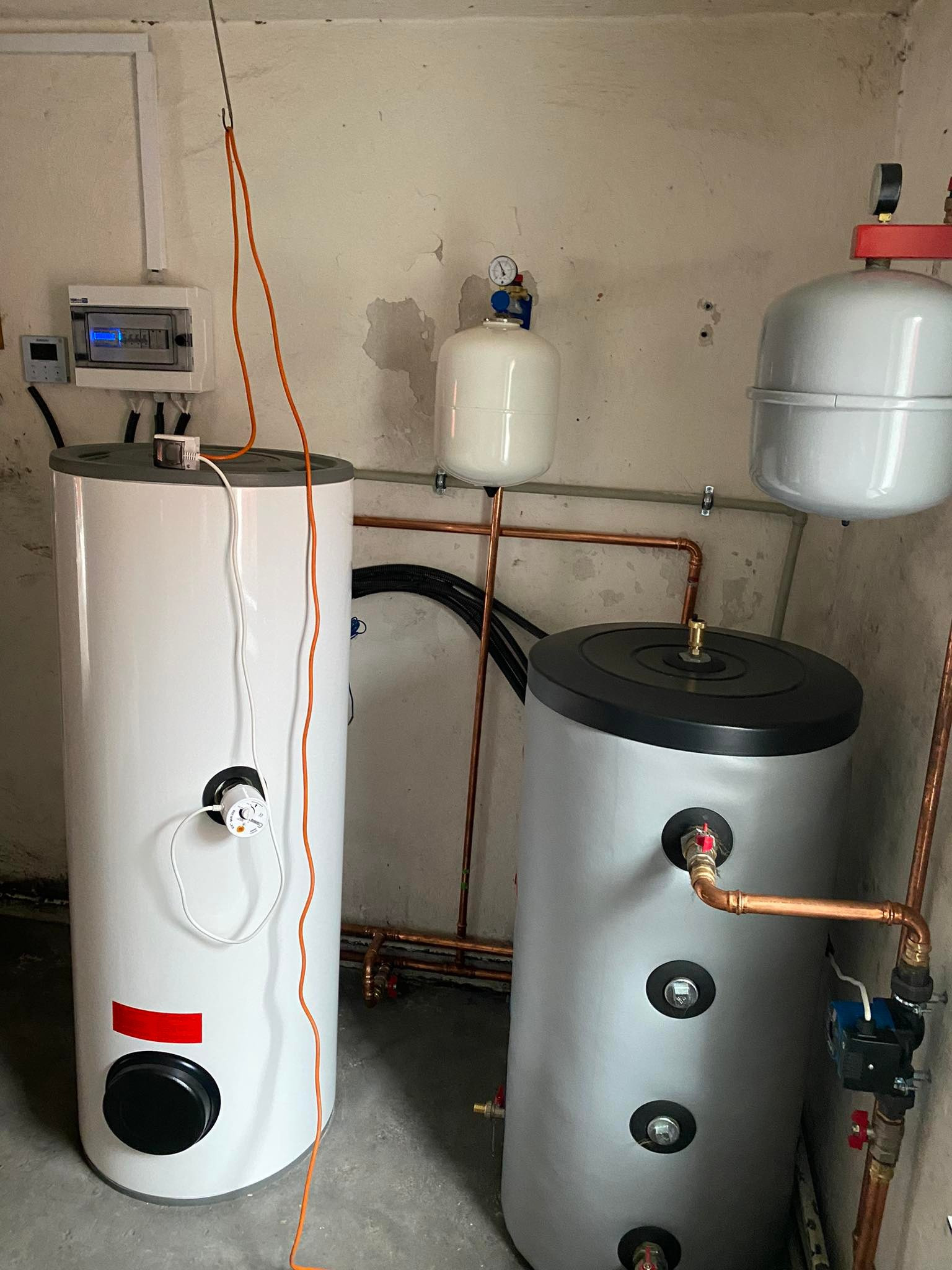 Dwa zasobniki ciepłej wody użytkowej: biały z czerwonym paskiem i szary z czarną górą, połączone miedzianymi rurami, z widocznymi elementami instalacji elektrycznej i hydraulicznymi.