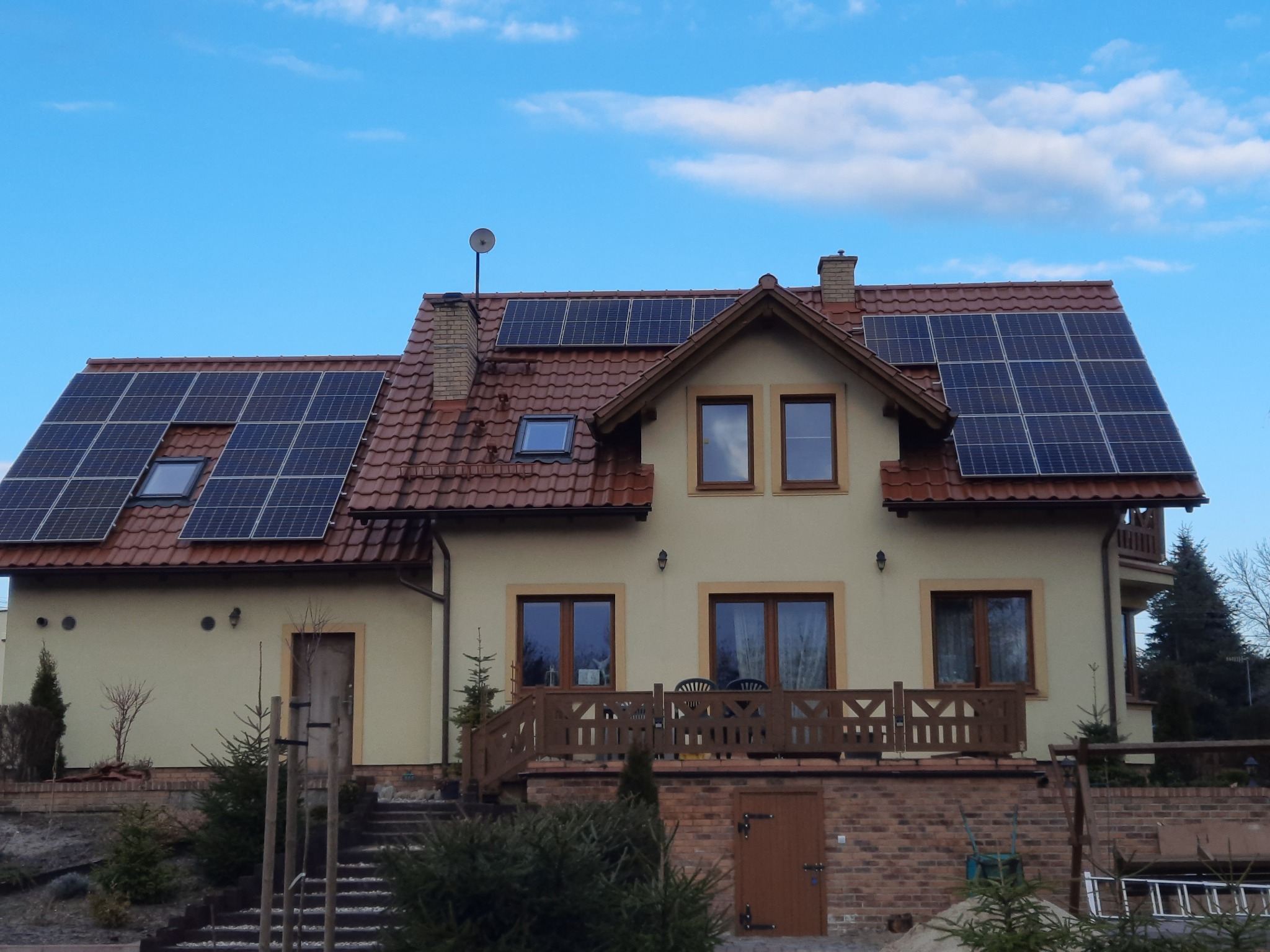 Dom jednorodzinny z panelami słonecznymi na dachu z czerwonej dachówki, antena satelitarna na kominie, jasne tło nieba.