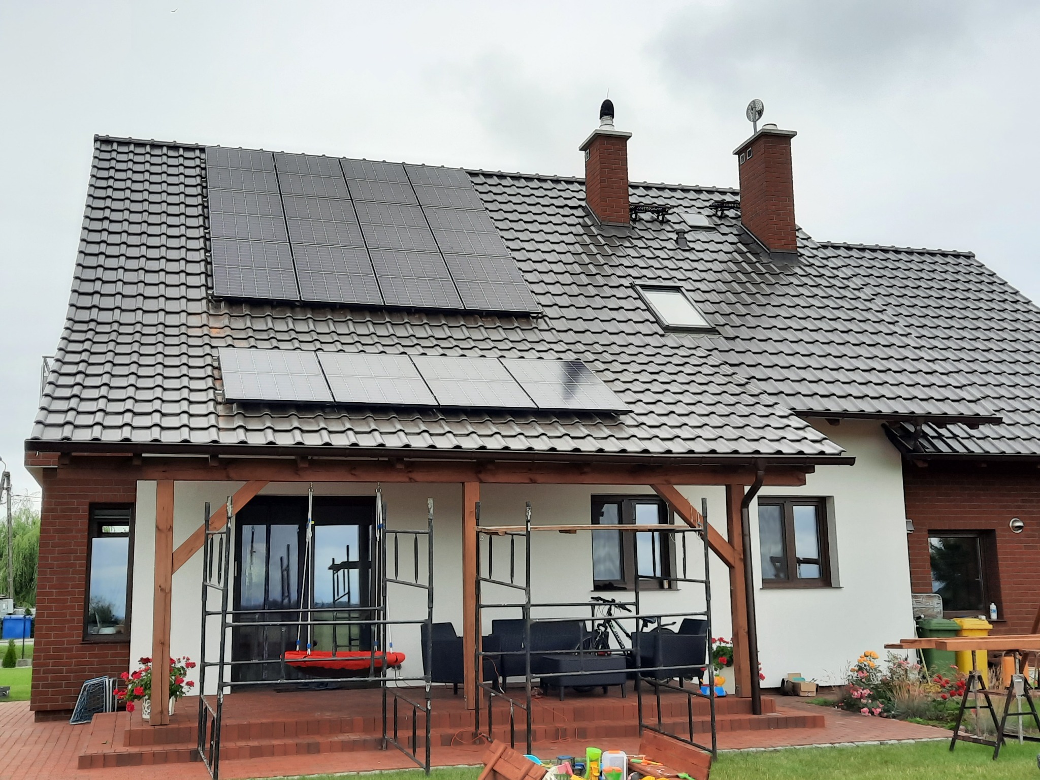 Instalacja paneli słonecznych na dachu domu jednorodzinnego z brązową dachówką, widoczne rusztowania i elementy montażowe na tarasie.