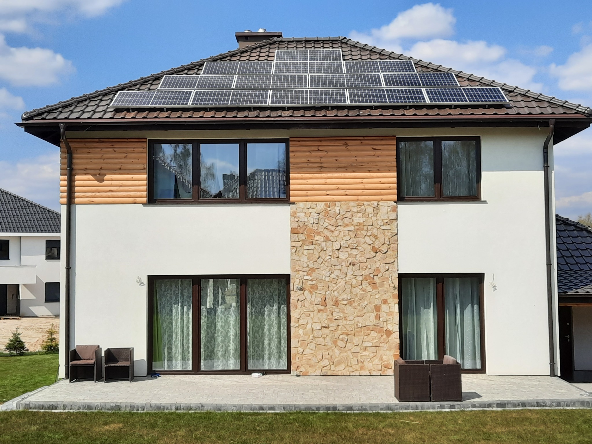 Dwukondygnacyjny dom jednorodzinny z panelami słonecznymi na dachu, elewacja z elementami drewnianymi i kamiennymi, widok z przodu na zadbany trawnik.