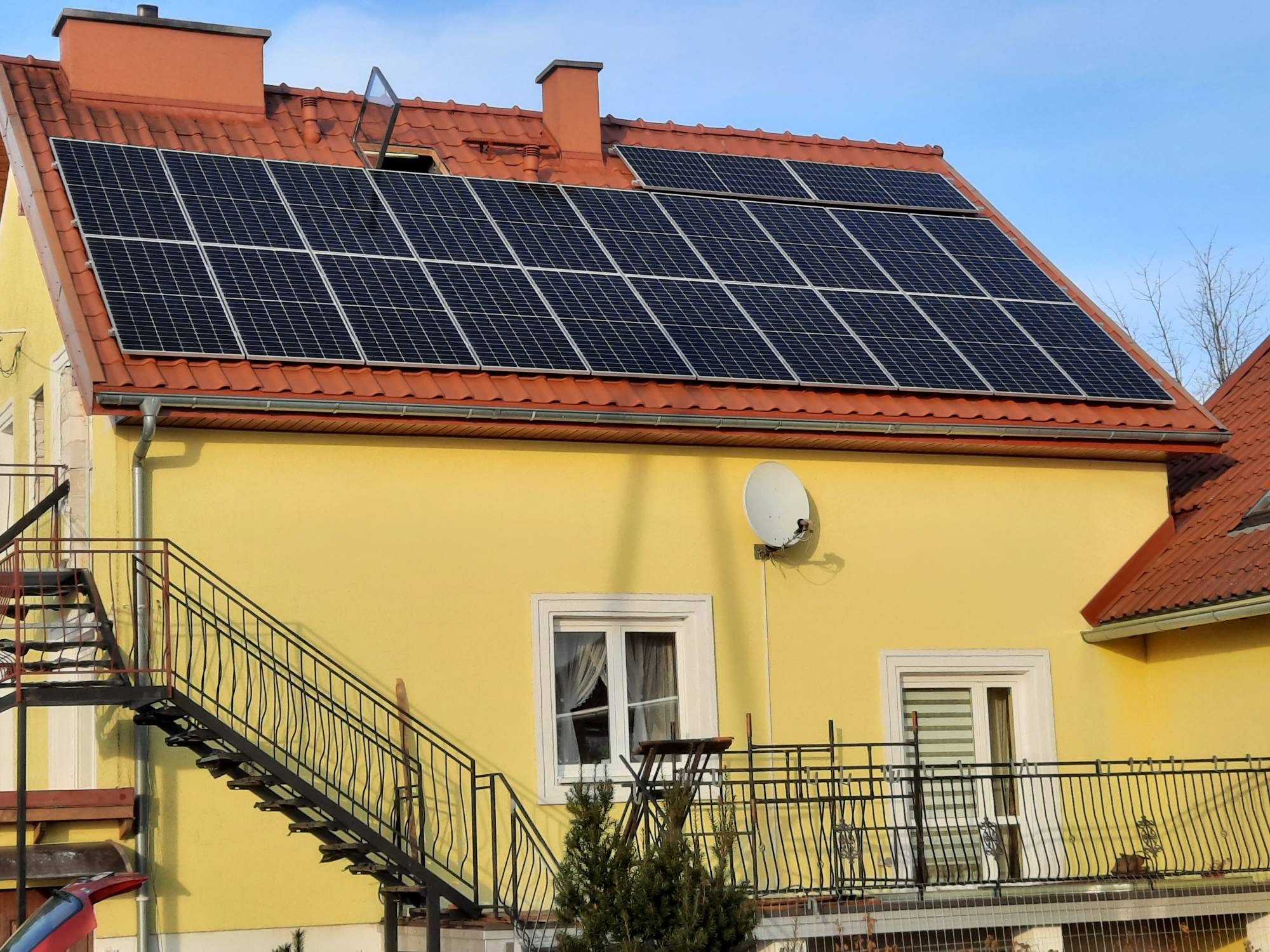 Żółty dom jednorodzinny z panelami słonecznymi na czerwonym dachu i zewnętrznymi schodami.