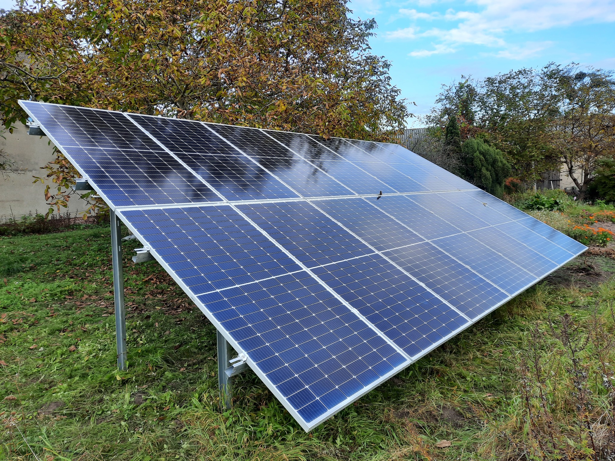 Panel słoneczny zamontowany na metalowej konstrukcji, otoczony zielenią trawy i drzew z jesiennymi liśćmi w tle, pod błękitnym niebem.