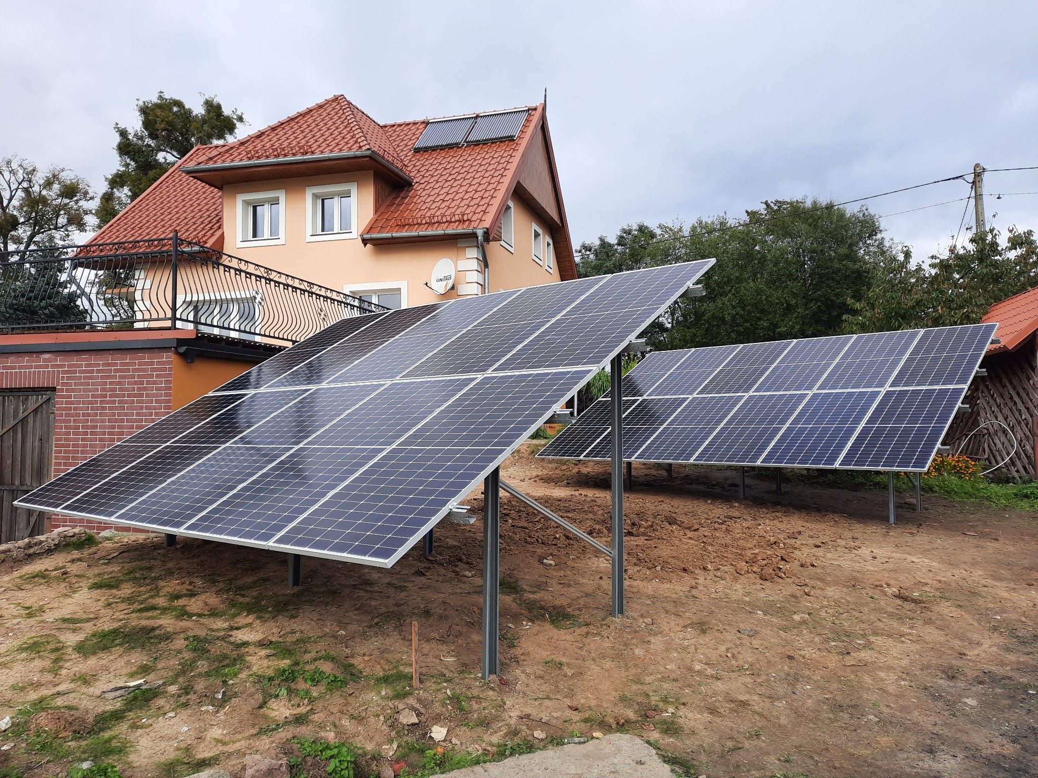 Dwa rzędy paneli słonecznych ustawione na ziemi przed domem z czerwoną dachówką i panelami słonecznymi na dachu. Widoczny kabel Unitron na elewacji budynku.