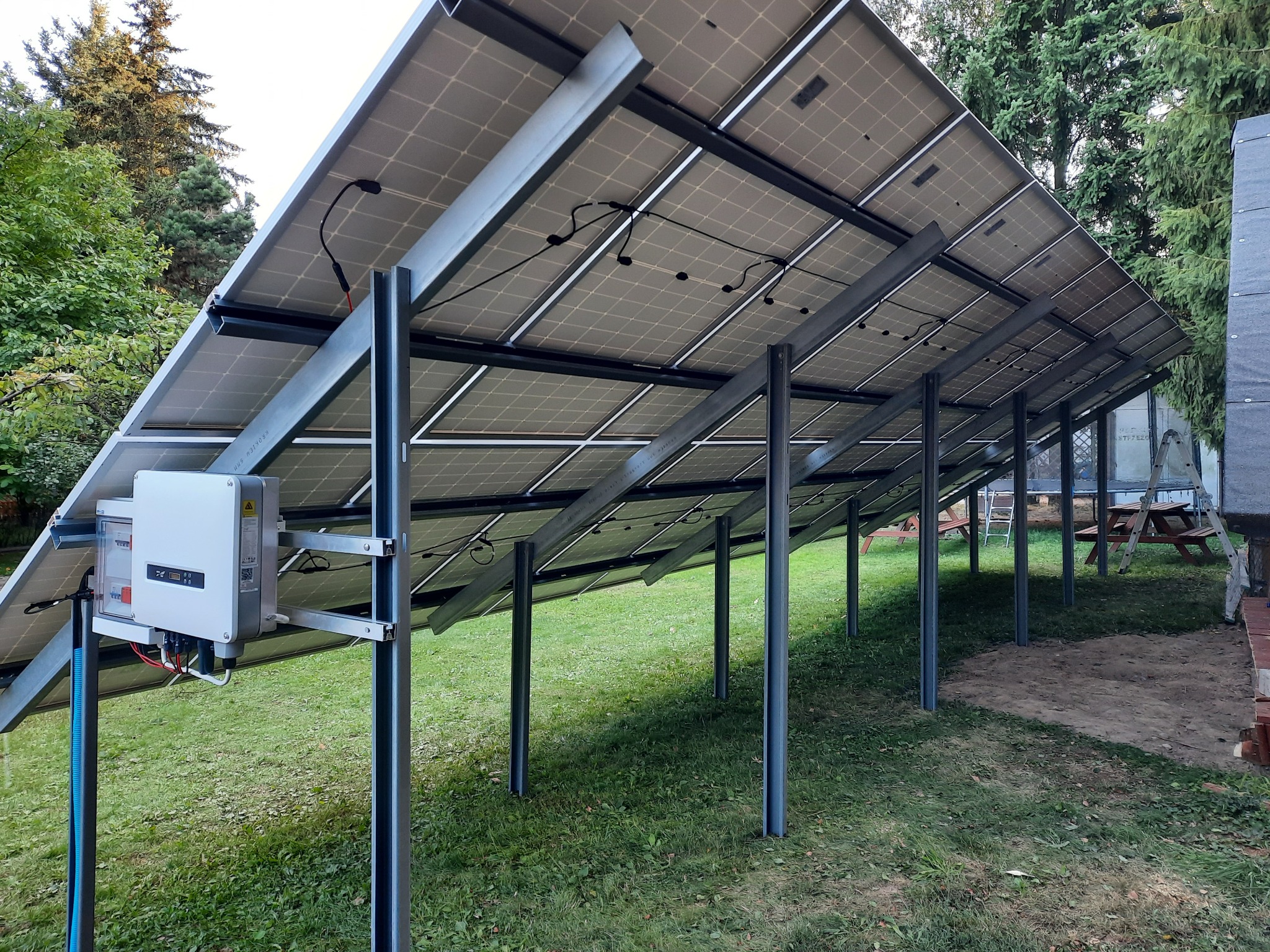 Widok od spodu na panele słoneczne zamontowane na metalowej konstrukcji na trawniku, widoczny inwerter, okablowanie i słupy podtrzymujące.