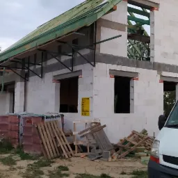 Budowa domu murowanego z białych bloczków, widoczne niezadaszone okna, drewniana konstrukcja dachu, komin w surowym stanie i materiały budowlane na placu budowy, częściowo zasłonięte przez...