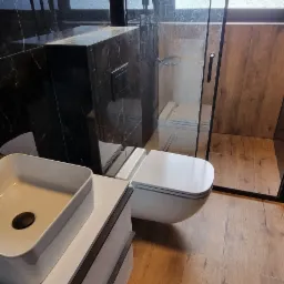 Nowoczesna łazienka z kabiną prysznicową typu walk-in, podwieszaną toaletą i umywalką nablatową na białej szafce, wykończona płytkami imitującymi drewno i czarnym marmurem.