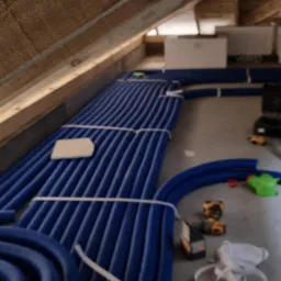 Niebieskie rury instalacyjne ułożone równolegle w przestrzeni poddasza, zabezpieczone białymi taśmami, widoczne narzędzia i elementy instalacji w tle.