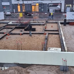 Instal-House Jankowski Dariusz - Fundamenty domu jednorodzinnego w trakcie budowy: widoczne izolowane ściany fundamentowe, zbrojenie pionowe, rury kanalizacyjne w piasku, teren budowy zabezpieczony ogrodzeniem tymczasowym.