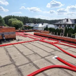Czerwone rury instalacji grzewczej, prawdopodobnie podłogowej, ułożone na szarym, płaskim dachu budynku w trakcie budowy, z widocznymi ceglanymi elementami i fragmentami instalacji.