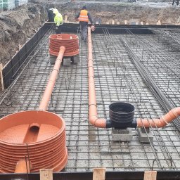 Instal-House Jankowski Dariusz - Pomarańczowe rury kanalizacyjne z tworzywa sztucznego, częściowo zamontowane w siatce zbrojeniowej fundamentu, widok z góry na placu budowy.
