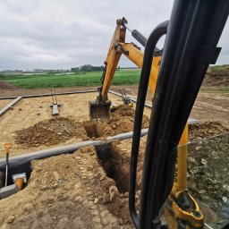 Instal-House Jankowski Dariusz - Żółta koparka wykopuje rów pod instalację na placu budowy z widokiem na pola, widoczne elementy instalacji, szpadel i zagęszczarka w tle.